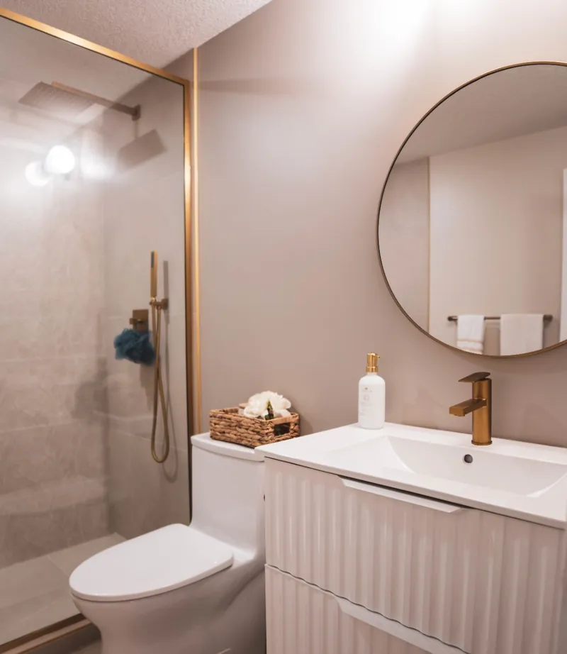 Basement bathroom with a glass-walled shower, a round mirror and a white fluted vanity with brass fixtures