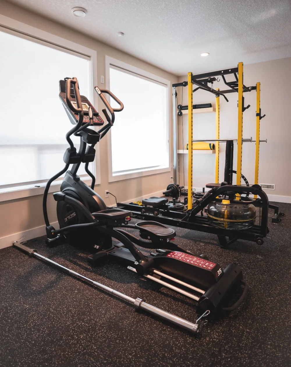 Basement home gym with rubber flooring, a power rack, an elliptical trainer and two large windows